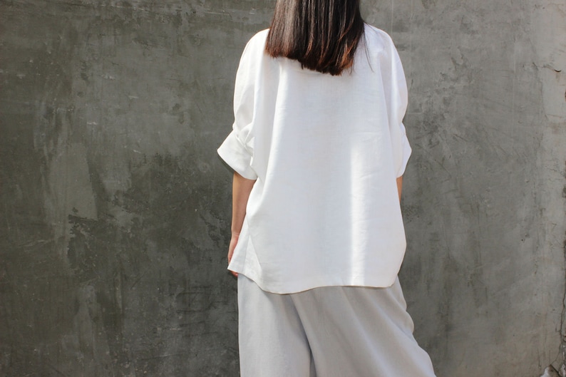 May include: A white linen shirt with a relaxed fit and three-quarter sleeves. The shirt is worn over a pair of light gray linen pants.