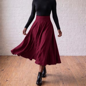 May include: A long, deep burgundy skirt with a high-waisted, flared silhouette that reaches below the knees. The skirt is paired with a long-sleeved black top and black leather ankle boots, creating a simple, solid-coloured ensemble.