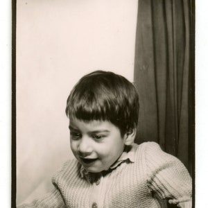 May include: A black and white photo of a young boy wearing a knitted sweater. He is smiling and looking to the side. The photo is taken in a studio setting with a curtain in the background.