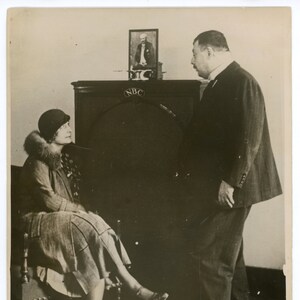 Op de afbeelding: Een zwart-witfoto van een man en een vrouw die voor een grote radio staan. De radio heeft de letters "NBC" erop. De vrouw draagt een hoed en een bontkraag. De man draagt een pak en een stropdas.