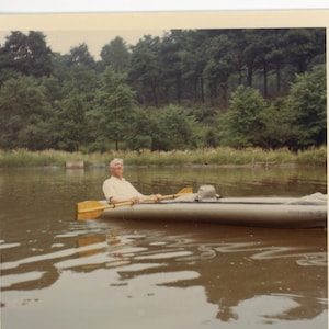 May include: A man in a white shirt paddles a canoe on a calm lake. The lake is surrounded by trees and greenery. The date "AUG 69" is written in the bottom right corner of the image.