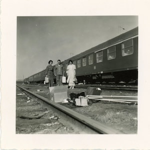 Può includere: Fotografia vintage in bianco e nero di tre persone su un binario accanto a un treno. Tengono bagagli, suggerendo che stanno per viaggiare. Le carrozze del treno hanno finestre rettangolari.