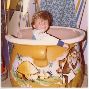 Könnte beinhalten: Ein kleines Kind sitzt in einer gelben, cartoonartigen Fahrt. Die Fahrt ist wie ein großer Topf mit einem Griff geformt und zeigt einen Cartoon-Affen mit einem Kochhut. Die Fahrt hat eine braune, strohgedeckte Hütte und grüne Palmen, die an die Seite gemalt sind.