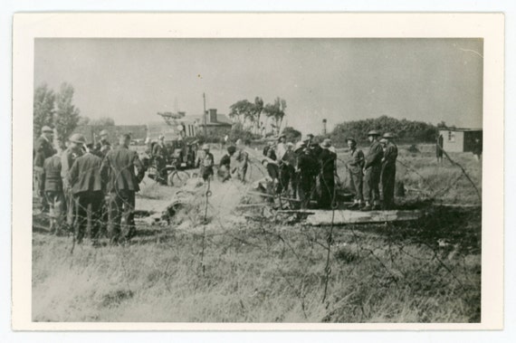 A group of men is gathered around the wreckage of a vintage airplane crash site, with some observing and others working nearby in an open field.