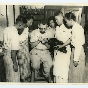 以下が含まれることがあります： 眼鏡をかけた男性を含む5人のグループが、白黒の動物の周りに集まっている。男性は動物を手に取り、じっと観察している。人々は白いシャツとチェック柄のスカートまたはズボンを着用している。シーンは、木製のキャビネットと棚のある部屋を背景にしている。