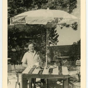 Può includere: Una fotografia in bianco e nero di un uomo seduto a un tavolo sotto un ombrello a righe. L'ombrello ha la scritta "Champagne" su di esso. L'uomo indossa una camicia di colore chiaro e tiene una tazza. Il tavolo è in legno e ha un motivo a scacchi.