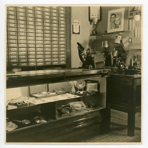 Könnte beinhalten: Ein Vintage-Optikergeschäft mit einer Glasvitrine gefüllt mit Brillenfassungen und Gläsern. Der Laden hat einen Holztheke mit einer Vielzahl von Werkzeugen und Geräten für Augenuntersuchungen und Reparaturen. An der Wand hängt ein gerahmtes Foto von zwei Personen.