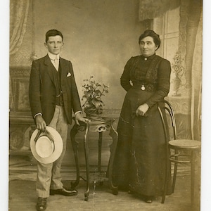 Puede incluir: Una fotografía antigua en blanco y negro de un joven y una joven de pie en una habitación. El hombre lleva un traje y sostiene un sombrero. La mujer lleva un vestido largo y está de pie junto a una silla. Hay una mesa pequeña con una planta encima en el fondo.