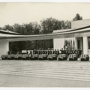 May include: A black and white photograph of a military funeral.  A row of caskets draped with American flags are lined up in front of a building.  Military personnel stand at attention in formation.