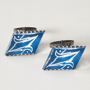 Op de afbeelding: Een paar manchetknopen met een blauw, ruitvormig ontwerp. Elke manchetknop heeft een wit, gestileerd ontwerp op een blauwe achtergrond, met een zilverkleurig metalen frame en sluiting.