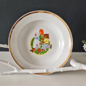 May include: A white ceramic bowl with a gold rim features a cartoon illustration of two bears, one holding a red balloon, walking on a green background.