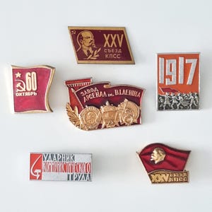 May include: A collection of six vintage enamel badges featuring red, gold and white colours. The badges depict various Soviet-era symbols and text, including "60 Октябрь" (60 October), "XXV съезд КПСС" (XXV Congress of the CPSU), "Завод Арсенал им. В.И. Ленина" (Arsenal Factory named after V.I. Lenin), "1917", and "Ударник Коммунистического Труда" (Shock Worker of Communist Labor).