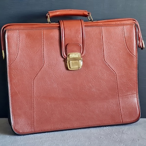 May include: A vintage brown briefcase with a handle and a gold-colored clasp. The briefcase has a textured surface and is likely made of leather or a leather-like material. The briefcase is closed and appears to be in good condition.