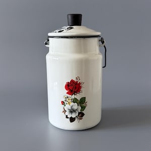 May include: A white enamel milk churn with a black handle and lid. The lid has a black knob and black floral detailing. A red rose and white flower bouquet decorates the front.