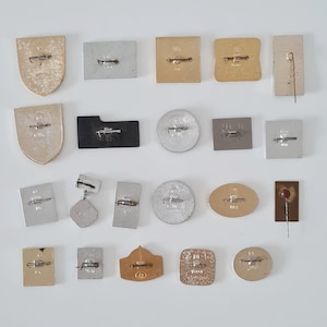 May include: A collection of nineteen metal pins and badges arranged in four rows, each featuring a horizontal pin-back mechanism on the reverse side. The items vary in shape, including shields, rectangles, circles, and ovals, with gold and silver tones.