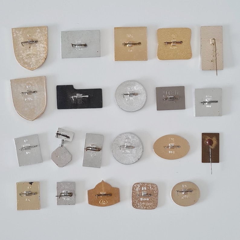 May include: A collection of nineteen metal pins and badges arranged in four rows, each featuring a horizontal pin-back mechanism on the reverse side. The items vary in shape, including shields, rectangles, circles, and ovals, with gold and silver tones.