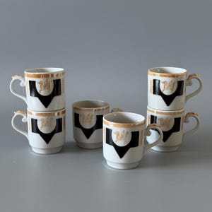 May include: Set of six white ceramic mugs with gold trim and handles. Each mug features a black and gold crest design. The mugs are stacked in pairs and arranged on a gray surface.