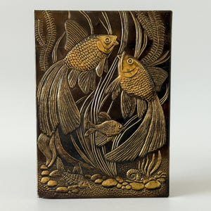 Puede incluir: Panel decorativo rectangular de color bronce con un diseño en relieve de tres peces dorados nadando entre plantas acuáticas. Los peces tienen escamas texturizadas y aletas fluidas, sobre un fondo de algas y guijarros estilizados.