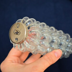 Puede incluir: Un tap&oacute;n de botella de vidrio transparente con una tapa redonda de lat&oacute;n. El tap&oacute;n de botella tiene forma de racimo de uvas.