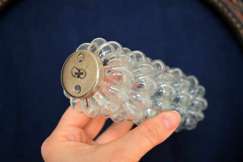 Puede incluir: Un tap&oacute;n de botella de vidrio transparente con una tapa redonda de lat&oacute;n. El tap&oacute;n de botella tiene forma de racimo de uvas.