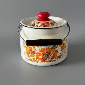 May include: A white enamel pot with a red lid and a dark brown handle. The pot is decorated with orange floral patterns. The pot has a metal handle and a lid with a red knob.