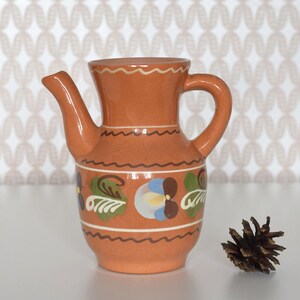 May include: A small, terracotta-coloured ceramic jug with a spout and handle. The jug is decorated with floral and leaf designs in blue, green, and white. A pine cone sits to the right of the jug.