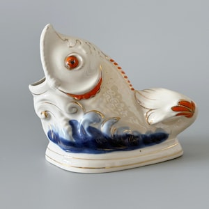 May include: A ceramic fish-shaped decorative piece. The fish is primarily white with orange accents on the mouth, fins, and eye. Blue waves are painted at the base, and gold trim outlines the edges. The fish has an open mouth and a detailed, textured surface.
