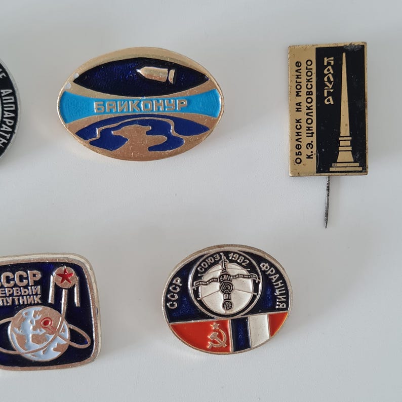 May include: A collection of four metal lapel pins featuring space exploration themes. One oval pin displays the text BAIKONUR, another rectangular pin shows a monument with Russian text, and two others depict Soviet space missions with Cyrillic text.
