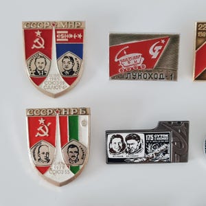 May include: A collection of four metal enamel pins featuring Soviet space exploration themes. Two shield-shaped pins display portraits of cosmonauts and mission text, while two rectangular pins depict a lunar rover and a space mission crew.