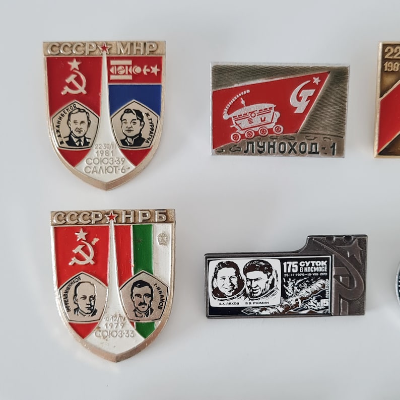 May include: A collection of four metal enamel pins featuring Soviet space exploration themes. Two shield-shaped pins display portraits of cosmonauts and mission text, while two rectangular pins depict a lunar rover and a space mission crew.