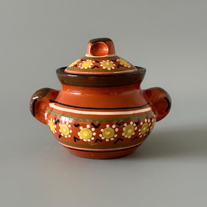 Könnte beinhalten: Eine weiße Keramik-Tasse mit schwarzem Henkel und dem Text "But First, Coffee" in fetter, schwarzer Schrift. Die Tasse steht vor einem verschwommenen Hintergrund. Die Tasse ist etwa 10 cm hoch.