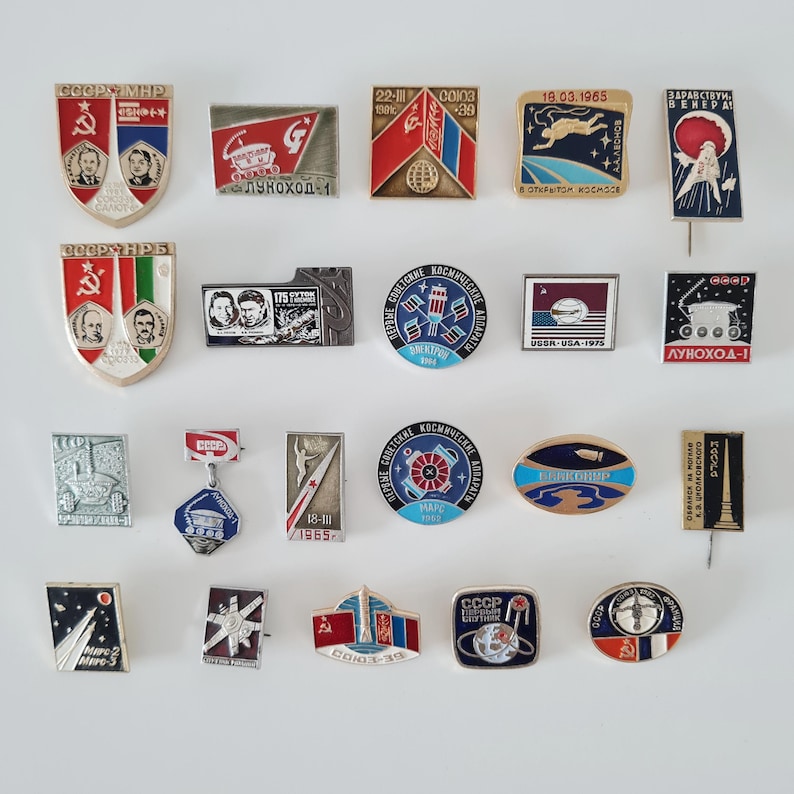 May include: A collection of nineteen vintage Soviet space-themed enamel pins featuring various shapes, including shields, rectangles, circles, and ovals. The pins display colorful graphics of rockets, cosmonauts, planets, and Cyrillic text related to space missions.