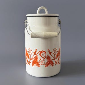 May include: Cream enamel milk churn with a lid and handle. The churn has a bright orange strawberry and floral pattern around the base. It has a metal handle and a wooden grip. The lid has a small handle for easy opening.