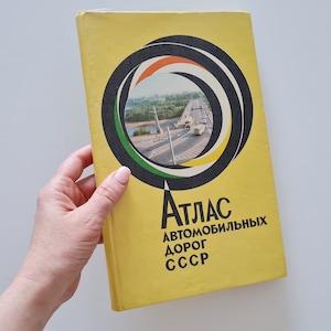 May include: A yellow book with a black and white circle design on the cover. The circle has a photo of a bridge and cars inside. The book title is "Atlas Avtomobilnykh Dorog SSSR".