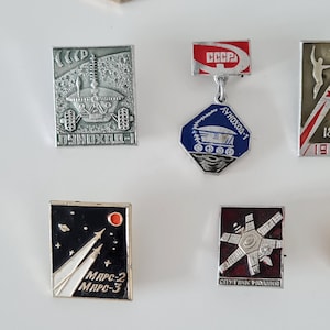 May include: A collection of five rectangular and hexagonal metal pins featuring Soviet space exploration themes. The pins display detailed silver-toned lunar rovers, rockets, and satellites against black, red, and blue enamel backgrounds with Cyrillic text.