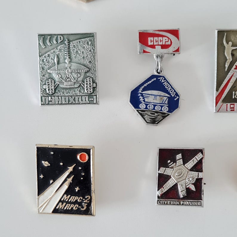 May include: A collection of five rectangular and hexagonal metal pins featuring Soviet space exploration themes. The pins display detailed silver-toned lunar rovers, rockets, and satellites against black, red, and blue enamel backgrounds with Cyrillic text.
