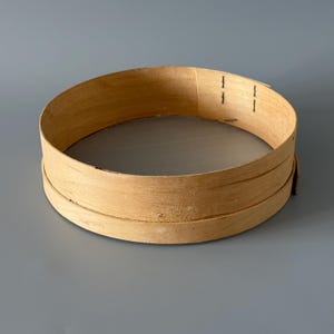 May include: A round wooden sieve featuring a light tan, thin bentwood frame with visible overlapping edges secured by two vertical staples. The base of the sieve consists of a fine, neutral-colored mesh screen stretched across the circular wooden rim.