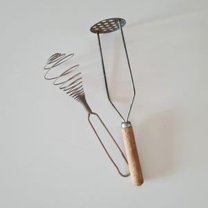 Può includere: Uno schiacciapatate in metallo con manico in legno e una frusta a filo. Lo schiacciapatate ha una testa circolare con fori distanziati. La frusta ha un design a spirale. Entrambi sono utensili da cucina.
