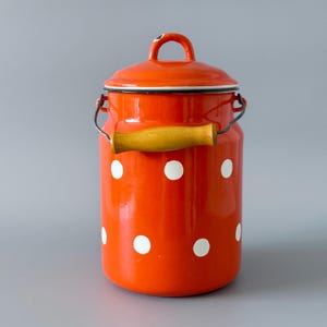 Op de afbeelding: Een feloranje geëmailleerde melkkan met een bijpassende deksel en houten handvat. De kan is versierd met witte stippen. De handgreep is van hout. De kan heeft een metalen draad handvat aan de zijkant.