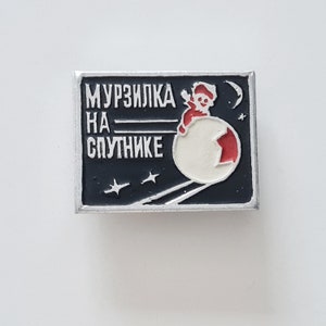 Puede incluir: Un pin de esmalte negro y blanco con una ilustración roja y blanca de un personaje de dibujos animados montando una esfera en el espacio. El texto "МУРЗИЛКА НА СПУТНИКЕ" está impreso en el pin.