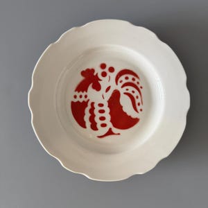 May include: A white ceramic plate with a scalloped edge and a red rooster design in the centre. The rooster is stylized with a detailed silhouette. The plate is set against a grey background.
