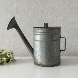 May include: A galvanized metal watering can with a long spout and a handle. The can has a cylindrical body with a lid and a small handle on the side. The watering can is designed for gardening and is made of durable metal.
