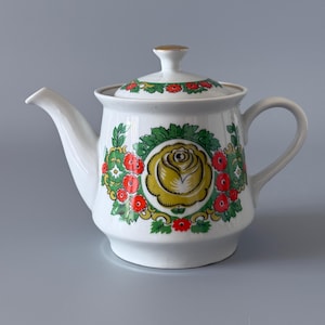 Könnte beinhalten: Weiße Keramik-Teekanne mit Deckel, verziert mit einem gelben Rosendesign, umgeben von roten und grünen floralen Akzenten. Die Teekanne hat einen gebogenen Ausguss, einen Henkel und einen goldfarbenen Knopf auf dem Deckel.