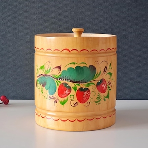 Pode incluir: Um recipiente de madeira com tampa, com morangos e folhas pintados à mão em vermelho, verde e amarelo. O pote cilíndrico tem um acabamento em madeira clara e detalhes vermelhos.