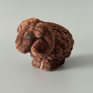 May include: A brown, plush ram figurine with detailed fur texture. The ram has curled horns, a small eye, and is posed in a standing position. The figurine is a decorative item.