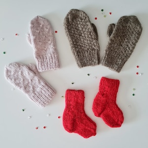 May include: A selection of hand-knitted winter accessories, featuring two pairs of mittens in pale pink and brown, and a pair of small red socks. The mittens have a cable knit pattern. The items are displayed on a white surface with scattered confetti.
