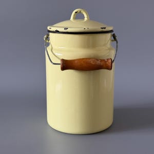 Könnte beinhalten: Eine hellgelbe Emaille-Milchkanne mit Deckel und Holzgriff. Die Kanne hat einen schwarzen Rand und einen Drahtgriff. Der Deckel hat einen kleinen Schlaufengriff. Die Kanne ist zylindrisch und hat eine Vintage-Ästhetik.