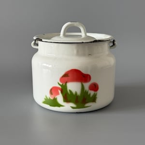 May include: A white enamelware jar with a lid and handle, featuring a red mushroom design with green grass. The jar has a cylindrical shape and small side handles. The lid has a loop handle.