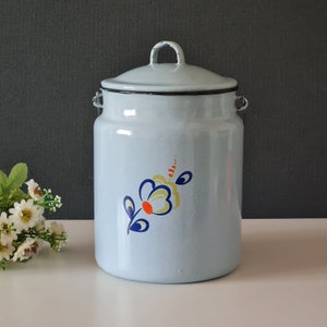 May include: A light blue enamel milk can with a floral design. The flower is yellow with a red center and blue leaves. The can has a lid and a handle.