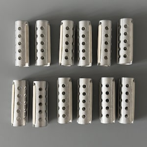 Peut inclure: Douze bigoudis argentés sont présentés. Chaque bigoudi est cylindrique, avec de multiples perforations circulaires et un bord plié. Les bigoudis sont disposés en trois rangées de quatre sur une surface grise.
